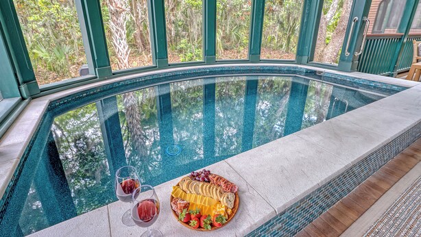 A pool with a glass edge, surrounded by greenery, and a table with a charcuterie board and drinks.