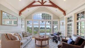 A spacious living room with a high ceiling, large windows, and a view of greenery outside.