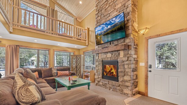 A spacious living room with a stone fireplace, a large flat-screen TV, and a balcony with wooden railings.