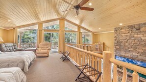 A spacious room with a high ceiling, wooden beams, a ceiling fan, large windows, a stone wall, and a balcony with a wooden railing.