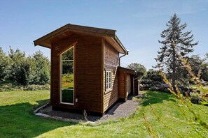 A wooden cabin with a large window and a door, surrounded by a grassy area and trees. - 4760 Aakirkeby - Sigtemøllevejen 11 (Aakirkeby)