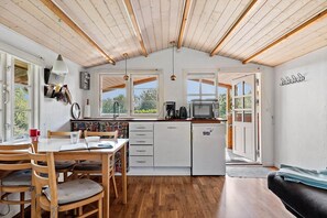 Une cuisine compacte avec des meubles en bois, une table à manger avec des chaises, et une vue sur l’extérieur par la porte ouverte.