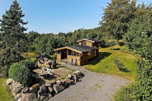 A wooden cabin with a patio area, surrounded by trees and a gravel path. - 4760 Aakirkeby - Sigtemøllevejen 11 (Aakirkeby)