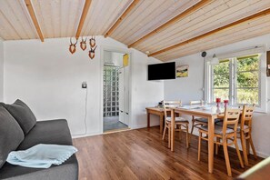 Ein Zimmer mit Holzboden, einem Esstisch mit Stühlen, einer Couch, einem an der Wand befestigten Fernseher und einem Fenster mit Blick auf Grünflächen.