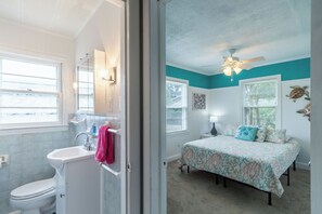 Ein Badezimmer mit Toilette, Waschbecken und Spiegel, sowie ein Schlafzimmer mit Bett, Fenstern und Deckenventilator.