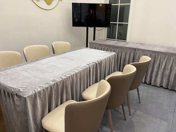 Un salón de conferencias con una larga mesa, sillas color beige, un televisor de pantalla plana y un ventanal con persianas.