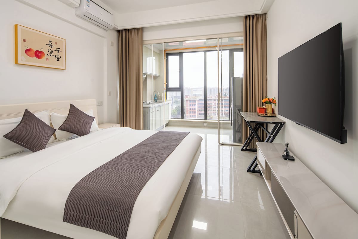 Quarto de hotel com uma cama grande, televisão de tela plana, uma mesinha com um vaso de flores e vista para a cidade pela janela.