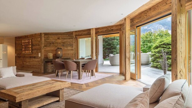 A modern interior with a wooden accent wall, a dining area with chairs, a sofa, and a large window offering a view of greenery.