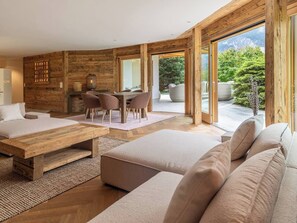 Ein modernes Interieur mit einer Wand in Holzoptik, einem Essbereich mit Stühlen, einer Couch und einem großen Fenster mit Blick ins Grüne.