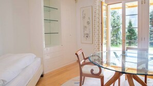 A bedroom with a bed, a glass table with chairs, a wardrobe, and a window with a view of trees.