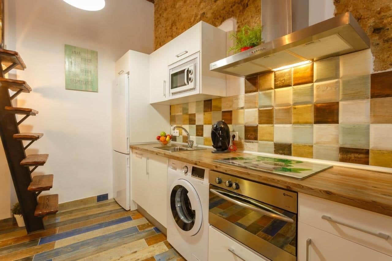 A compact kitchen with a built-in oven, microwave, and washing machine, a wooden staircase, and a checkered tile backsplash.