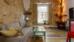 Una habitación rústica con paredes de piedra vista, suelo de madera, un sofá con cojines estampados, una mesa de vidrio, chimenea y televisión.