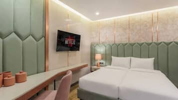 A hotel room with a bed, a desk, a chair, a TV mounted on the wall, and a lamp on the nightstand.