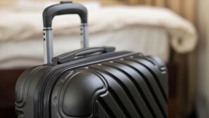 Une valise noire à roulettes, dotée d’une poignée et de roues, se trouve dans une chambre d’hôtel. On aperçoit un lit et des rideaux en arrière-plan.