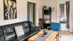 A living room with a black leather sofa, a wooden coffee table, and a blue armchair. There is a bookshelf with plants and a painting of an elephant. - 1 bedroom amazing apartment in Orrefors (Orrefors)