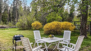 An outdoor seating area with a grill, white chairs, and a table with a flower arrangement, surrounded by trees and a yellow flowering bush. - 1 bedroom amazing apartment in Orrefors (Orrefors)