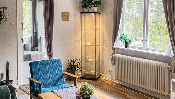 A cozy living room with a blue armchair, a glass cabinet with plants, a radiator, and a view of trees through the window. - 1 bedroom amazing apartment in Orrefors (Orrefors)