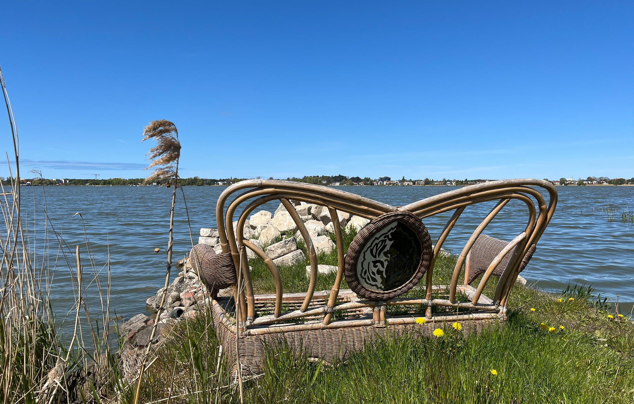 Une chaise en osier avec un panier tressé, placée sur une pelouse près d’une étendue d’eau.