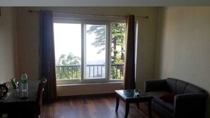 A living room with a sofa, a coffee table, and a dining table with chairs. There is a large window with curtains, and a view of trees and a balcony outside. - HOTEL PHOENIX (Mussoorie)