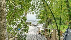 A wooden deck leading to a sandy area with a table and chairs, surrounded by lush greenery and a view of a body of water. - Private Beach + Outdoor Oasis: Home in St Pete! (St. Petersburg)