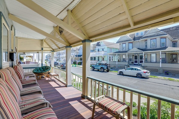 A covered porch with striped seating, a green chair, and a view of a street with parked cars and houses. - Walk to Beach & Dtwn: Apt in Ocean Grove (Ocean Grove)