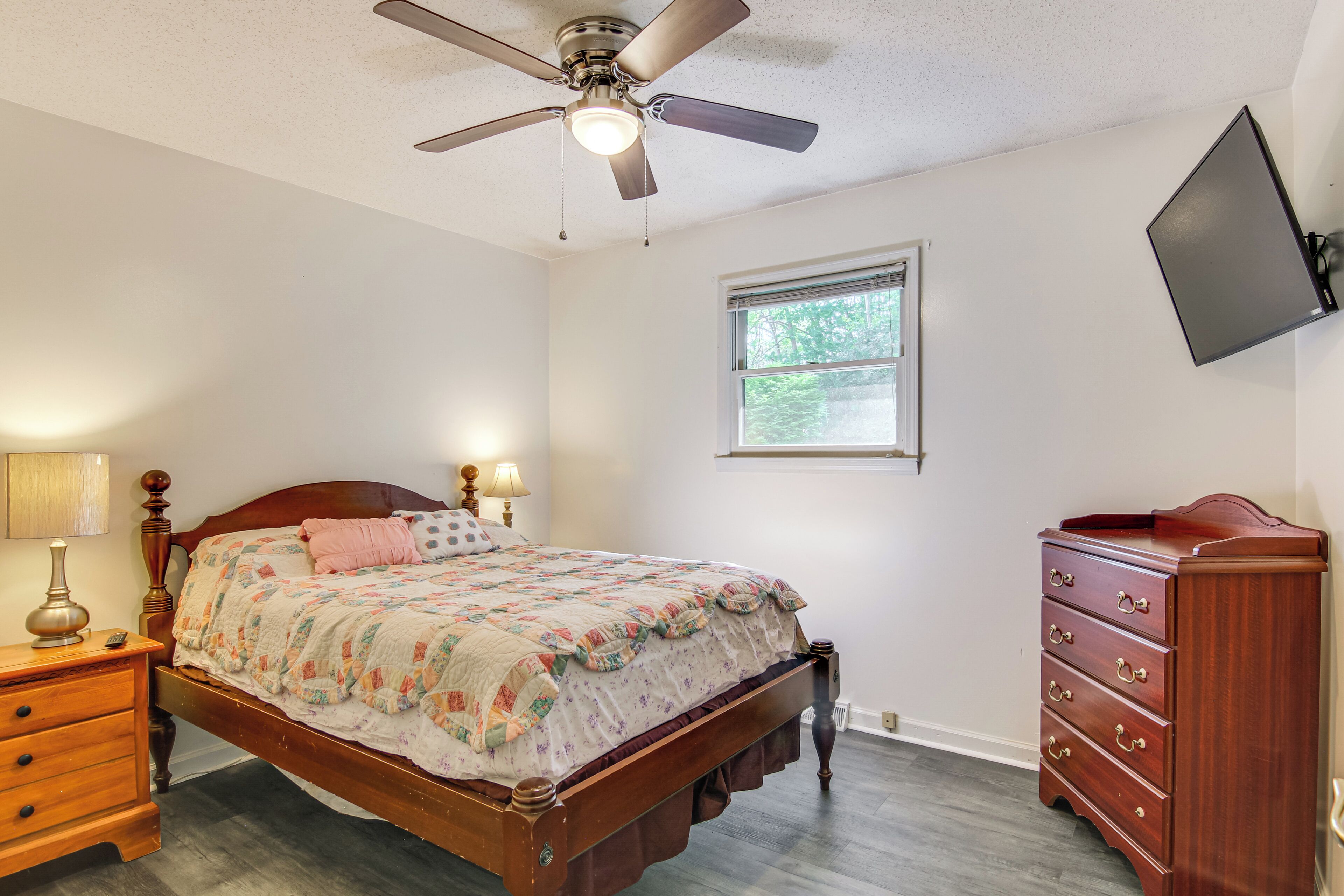 A bedroom with a wooden bed, a nightstand with a lamp, a dresser, a ceiling fan, and a window with a view of trees.