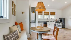Un espacio de sala acogedor con una mesa redonda de madera y sillas, un banco con cojines, una lámpara colgante de mimbre y una cocina al fondo.