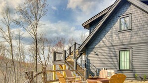 Una casa con una escalera que sube a una terraza, mobiliario exterior con sillas tipo Adirondack y un ventanal grande.