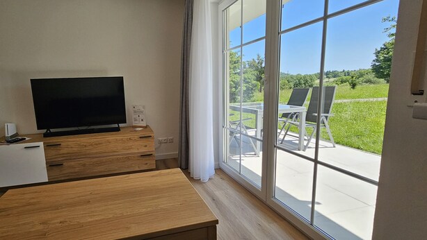 A modern room with a TV on a wooden stand, a small white cabinet, and a bench. Large windows offer a view of a green outdoor area with chairs. - Mountain idyll -renovated and high quality with solid wood bed at comfort height (Altenberg)