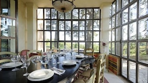 Un espace repas avec une longue table dressée pour le repas, de grandes fenêtres et un lustre.