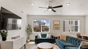 A modern living room with a large flat-screen TV, a ceiling fan, and a view of the outside buildings through large windows.