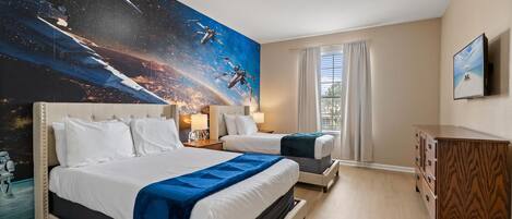 A hotel room with a large space-themed mural on the wall, two beds, a wooden dresser, and a television on the wall.