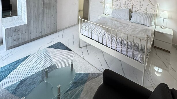 Un dormitorio moderno con piso de mármol, una cama con estructura metálica, una mesa de centro de vidrio y un armario con panel de madera.