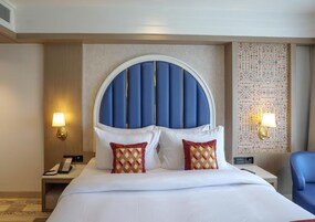 Una habitación de hotel con una cama grande, cabecero, dos lámparas de noche, mesitas de noche, un teléfono y una silla azul.