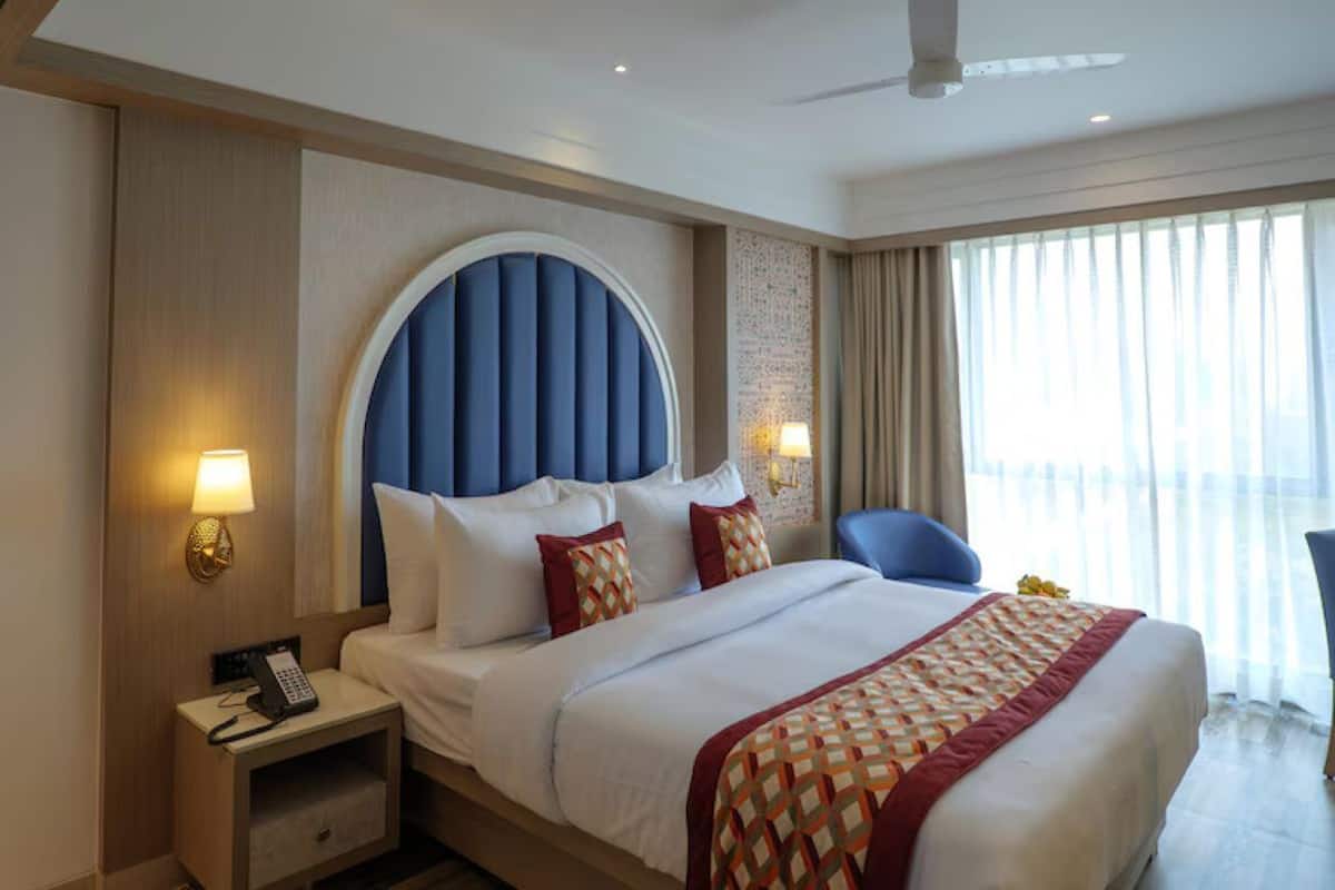 A hotel room with a large bed, a nightstand with a phone, and a view of the outdoors through large windows.