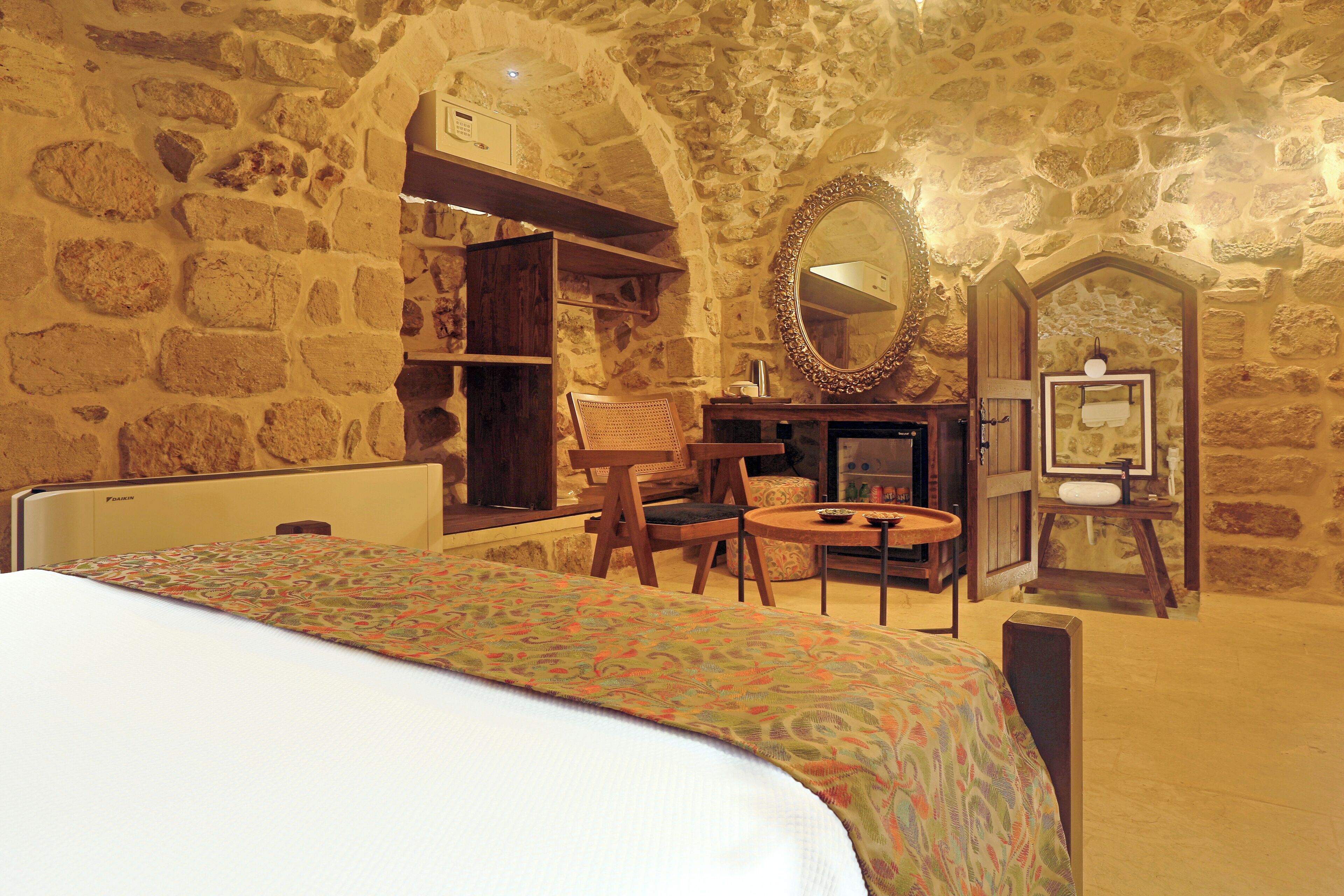 Une pièce comprenant un lit, une chaise, une table et un miroir. On y trouve également une armoire en bois et un mur en pierre.