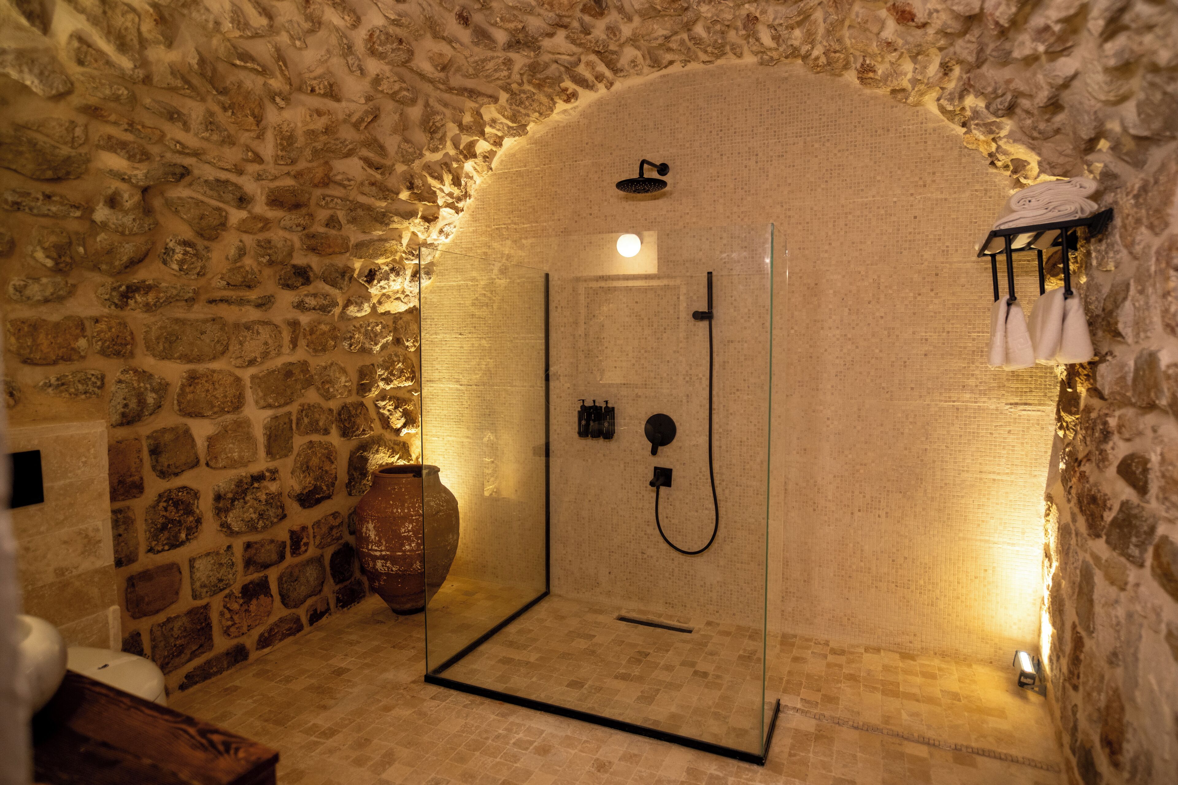 A stone-walled shower room with a glass door, a towel rack, and a large vase.