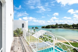 A balcony with a view of a clear blue sea, a wooden deck, and a white railing.