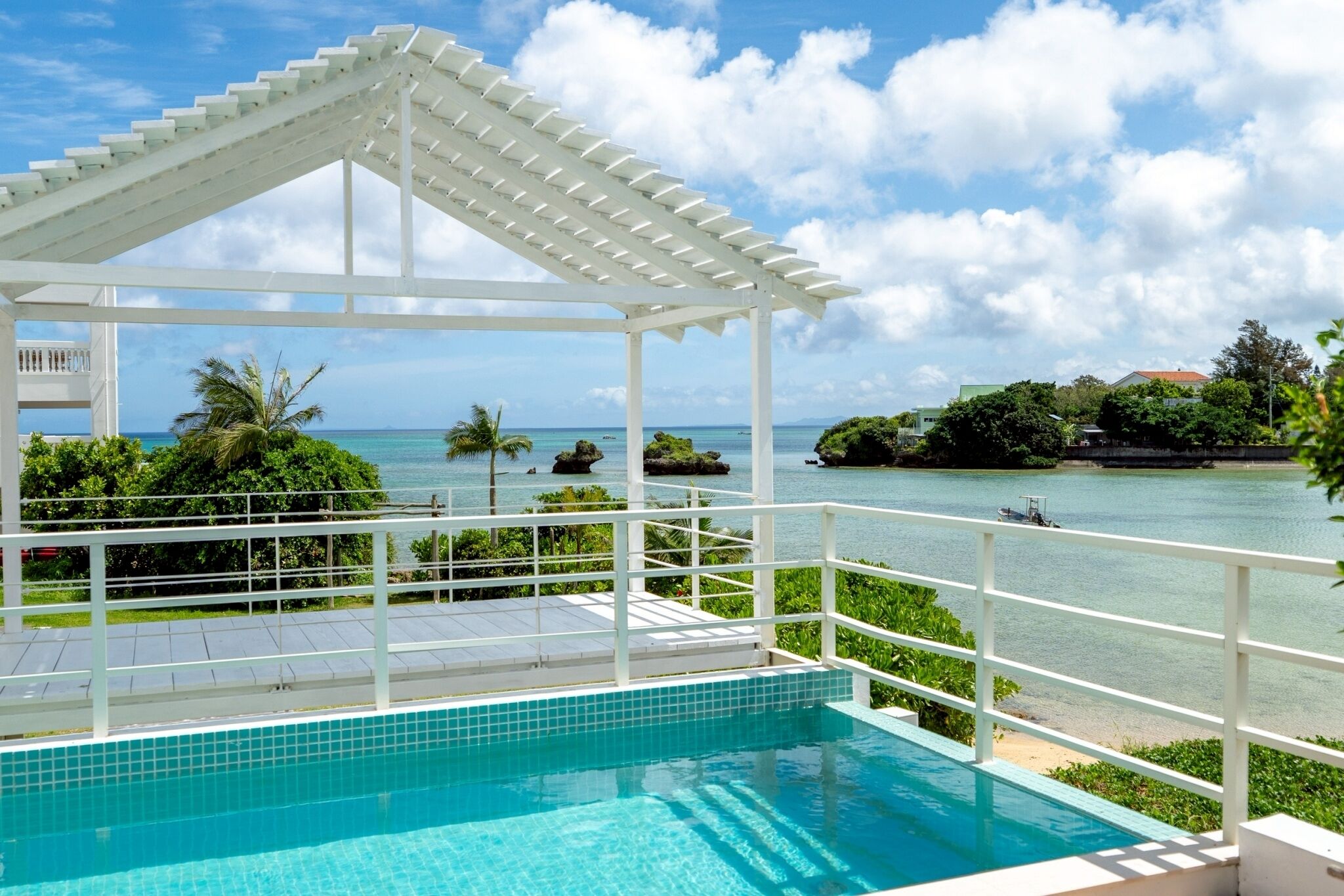 A pool with a white pergola, surrounded by lush greenery and a view of the sea.