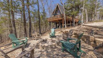 A cabin with a deck, surrounded by trees, and furnished with Adirondack chairs.