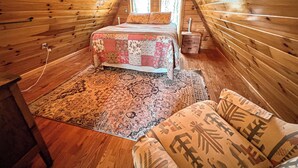 A cozy attic bedroom with wooden walls, a bed with a floral quilt, a patterned rug, and a small wooden nightstand.