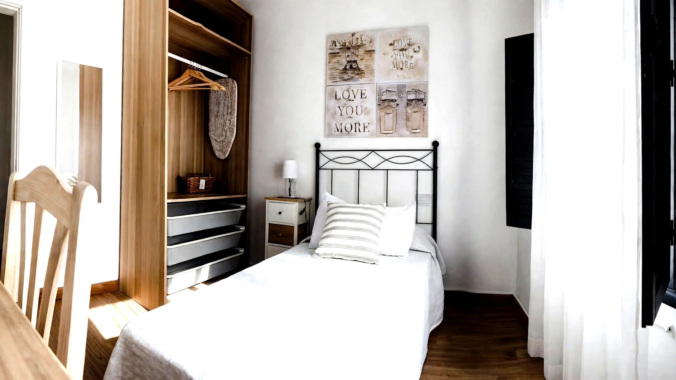 A bedroom with a bed, a nightstand, a wooden wardrobe, and a wall with artwork.