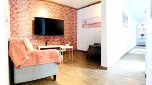 Una sala de estar con pared de ladrillo, un televisor de pantalla plana, un sofá gris, una mesita pequeña y un sillón oscuro.