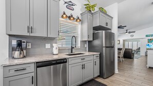 A modern kitchen with grey cabinets, a stainless steel refrigerator, a dishwasher, and a coffee maker. There is a window with blinds and a ceiling fan. - Dog Friendly and STEPS from the Ocean! Big Beach House with Pool, BBQ & Garage (New Smyrna Beach)