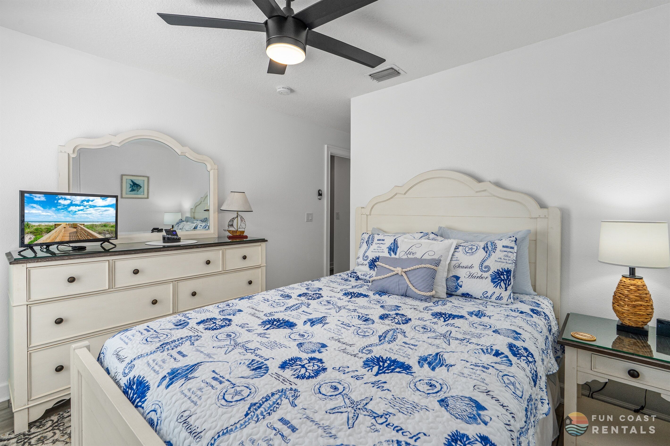 Un dormitorio con cama, ventilador de techo, mesita de noche, lámpara, cómoda, espejo y televisor.