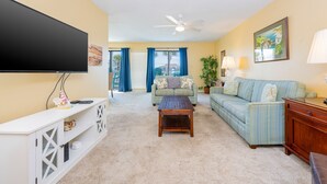 A living room with a flat-screen TV, a wooden coffee table, a striped sofa, and a white entertainment unit. - Sea Oats 105h 3 Bedroom Condo (Gulf Shores)
