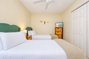 A bedroom with two beds, a ceiling fan, a television on a wooden stand, and a closet with a white door. - Sea Oats 105h 3 Bedroom Condo (Gulf Shores)