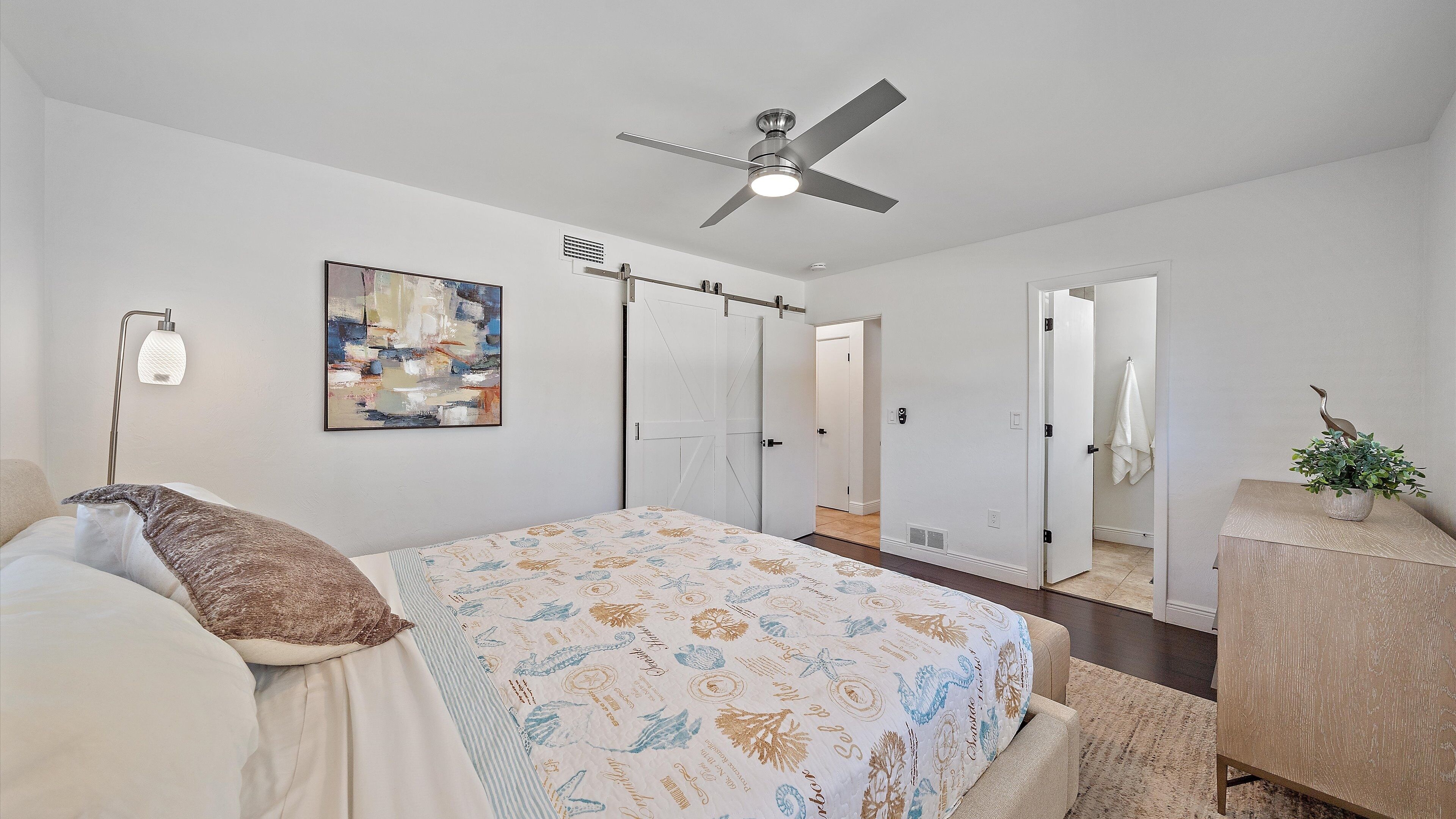 Une chambre à coucher avec un lit, un ventilateur de plafond, un tableau et une plante posée sur une table d’appoint.
