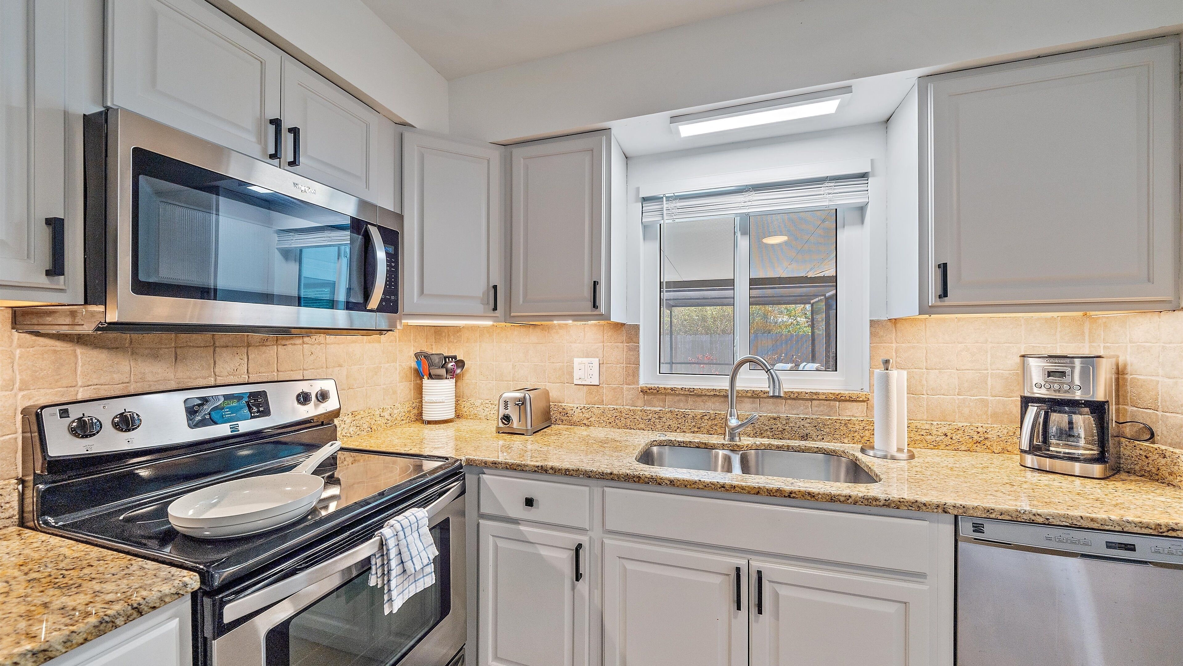 A modern kitchen with white cabinets, a stainless steel microwave, an oven, a sink, and a coffee maker.