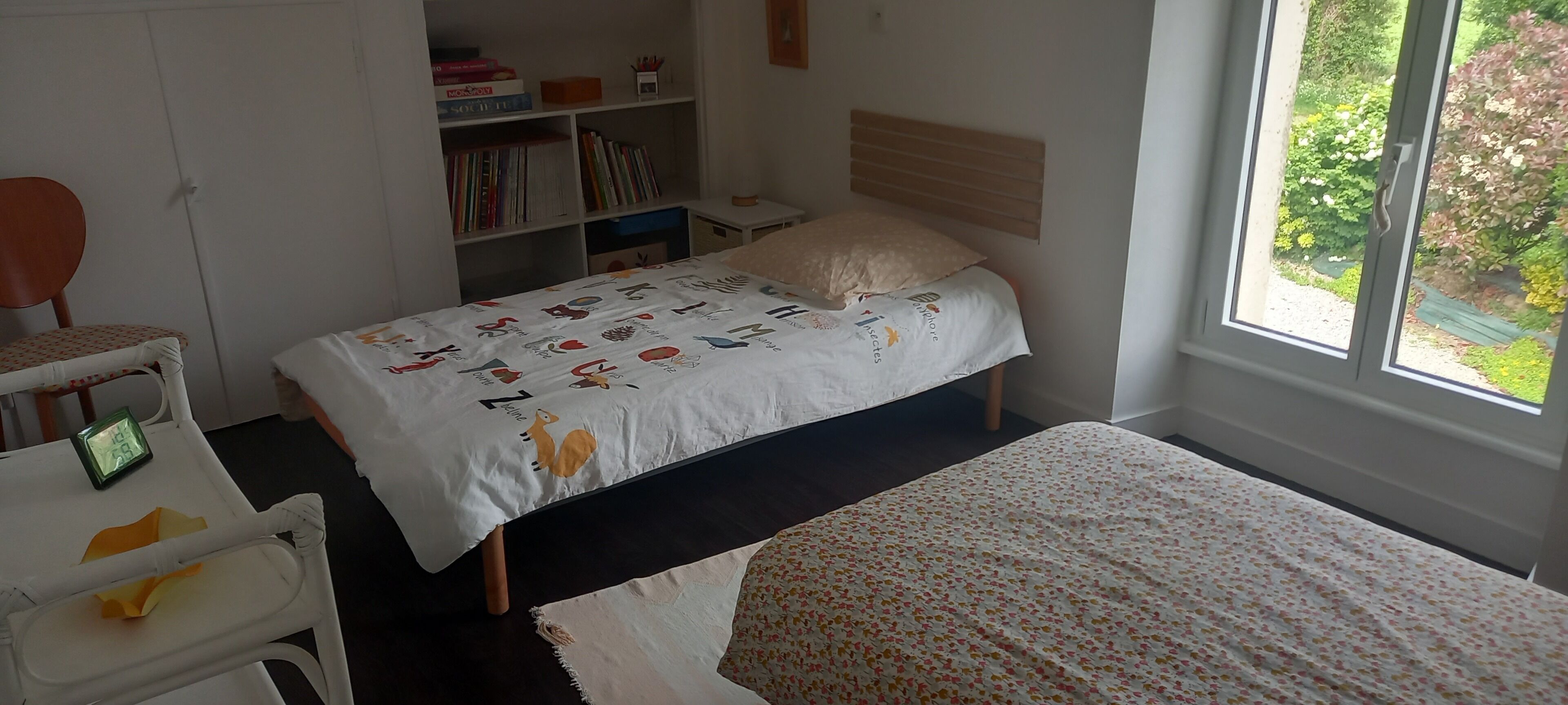 A bedroom with a bed, a chair, a small desk, a bookshelf, and a window with a view of greenery.
